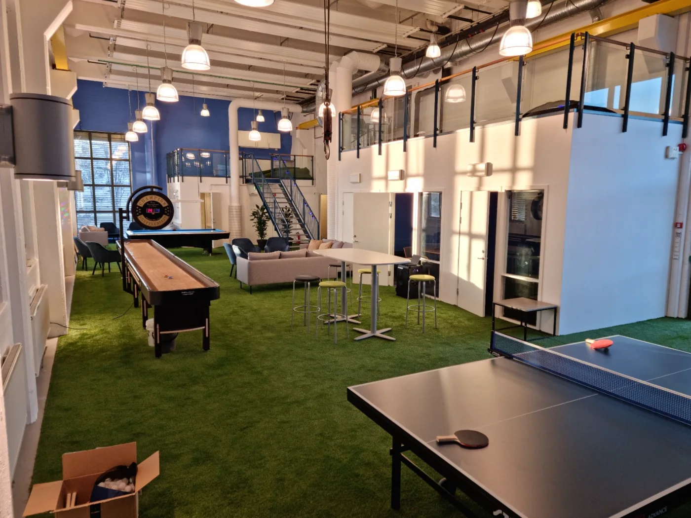 Shuffleboard, mesas altas e sofás distribuídos sobre a grama sintética
