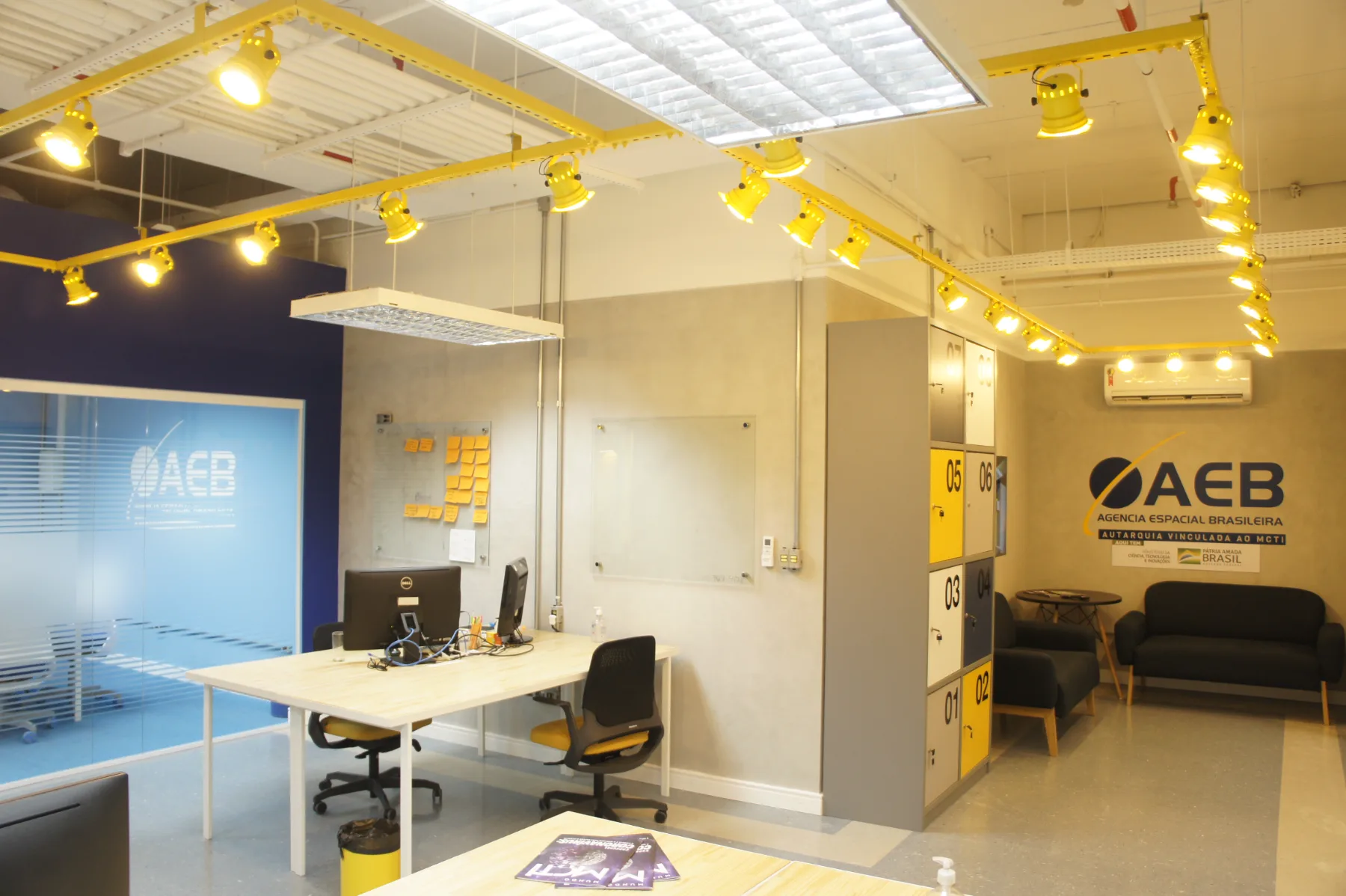 Unidade da AEB em open space: trilhos amarelos de iluminação sob a laje aparente, estações de trabalho compartilhadas, armários numerados e a marca aplicada na parede de cimento queimado