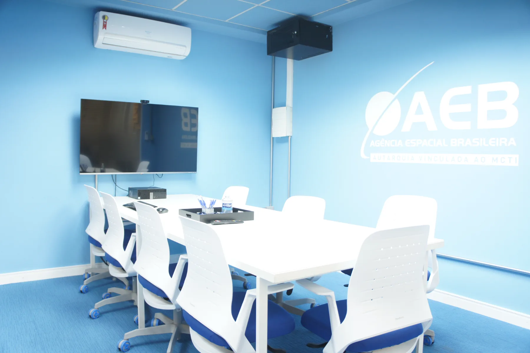 Interior da sala de reunião: mesa branca para oito lugares, monitor de videoconferência e a marca da AEB aplicada na parede azul
