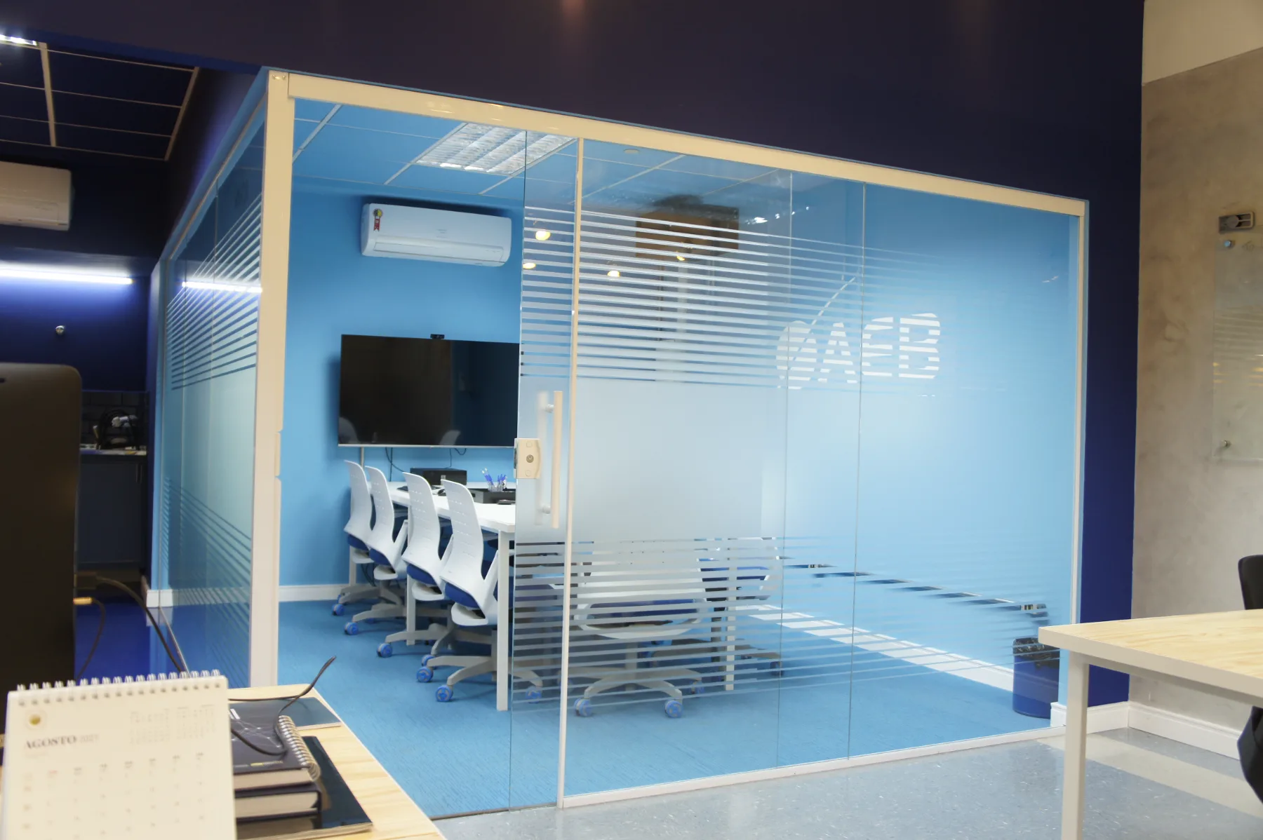 Sala de reunião em caixa de vidro com faixas jateadas e a marca da AEB, aberta para o open space de trabalho