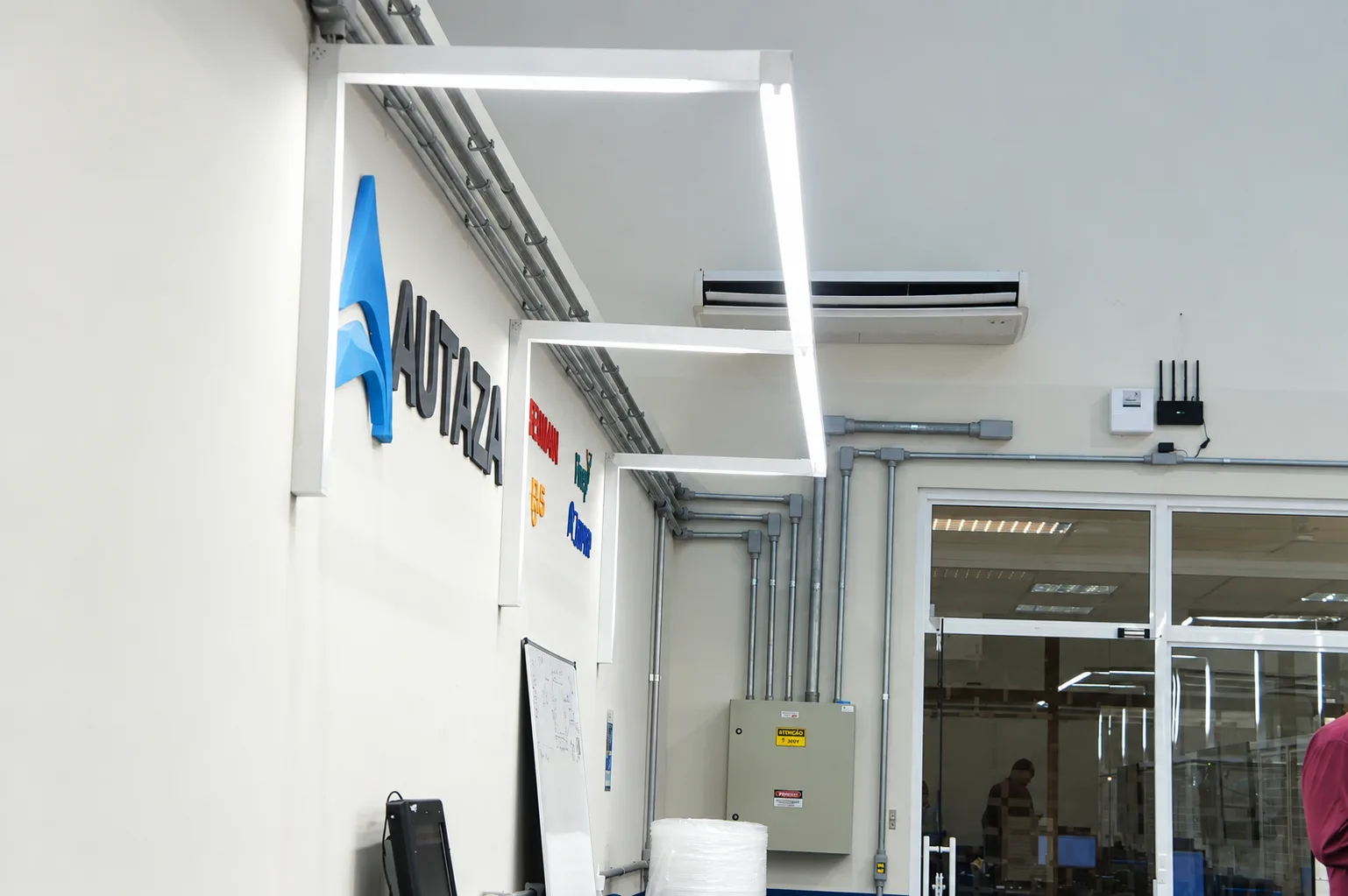 Marca da Autaza em relevo na parede do laboratório, ao lado das luminárias em pórtico e da infraestrutura elétrica aparente