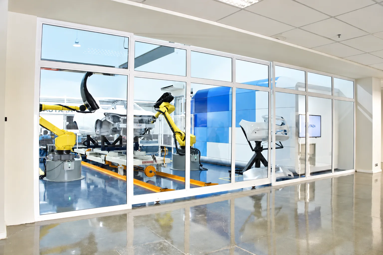 Laboratório visto do saguão através do painel envidraçado contínuo, com a célula robótica, o volume da sala óptica e o piso polido refletindo o conjunto