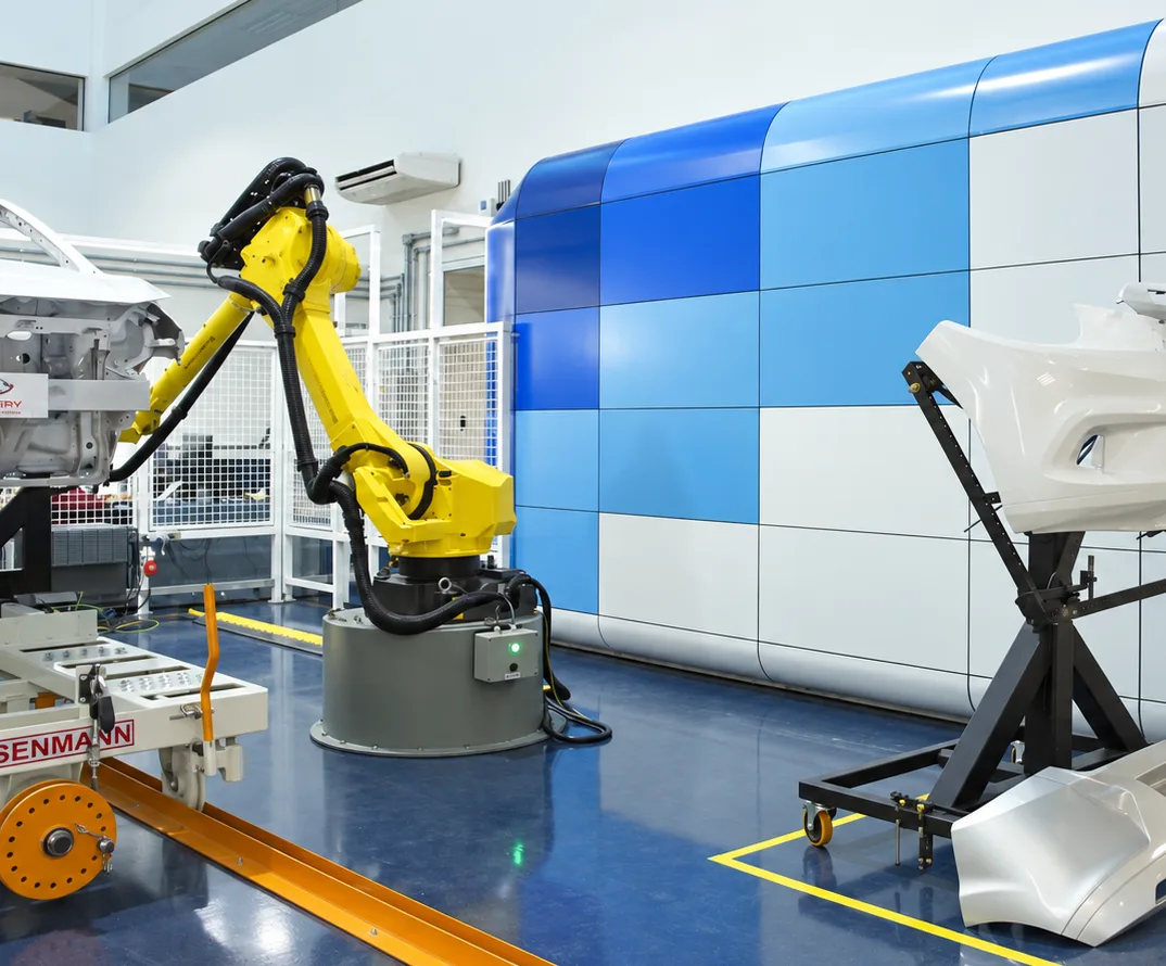 Braço robótico amarelo entre a carroceria e o para-choque em suporte móvel, diante do volume curvo em xadrez azul