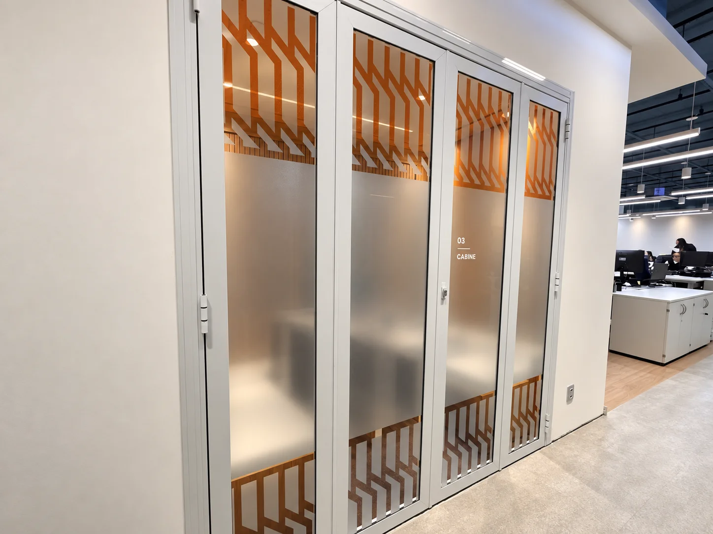 Cabine de chamada fechada no limite do open space: portas em vidro jateado com grafismo laranja e identificação numerada