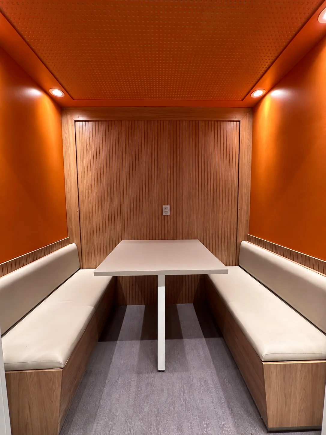 Interior da cabine: bancos estofados frente a frente, mesa branca, painel de madeira ripada e paredes laranja com forro acústico perfurado