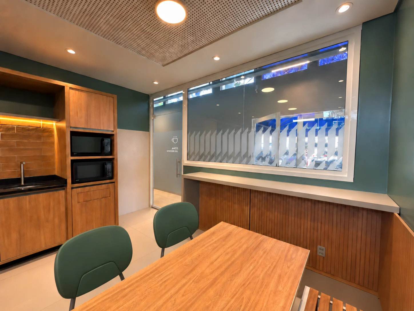 Interior da copa visto da mesa: bancada de apoio com micro-ondas, balcão sob a janela interna e vista para o open space
