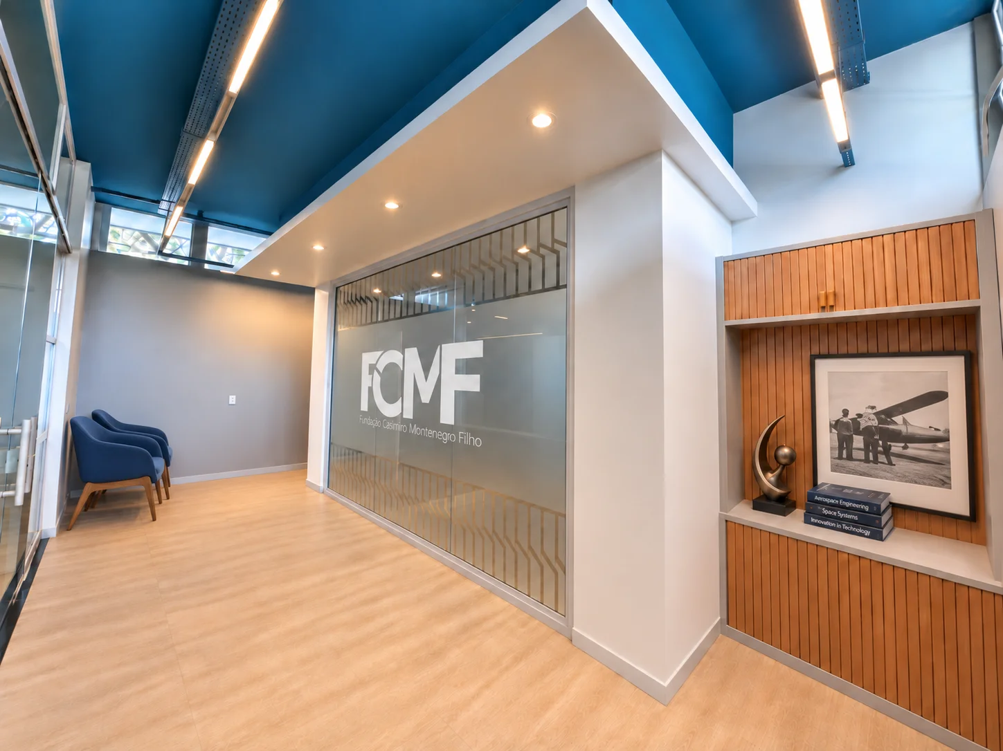 Lobby da sede da FCMF: forro azul com luminárias lineares, divisória de vidro com a marca da Fundação e nicho em madeira ripada