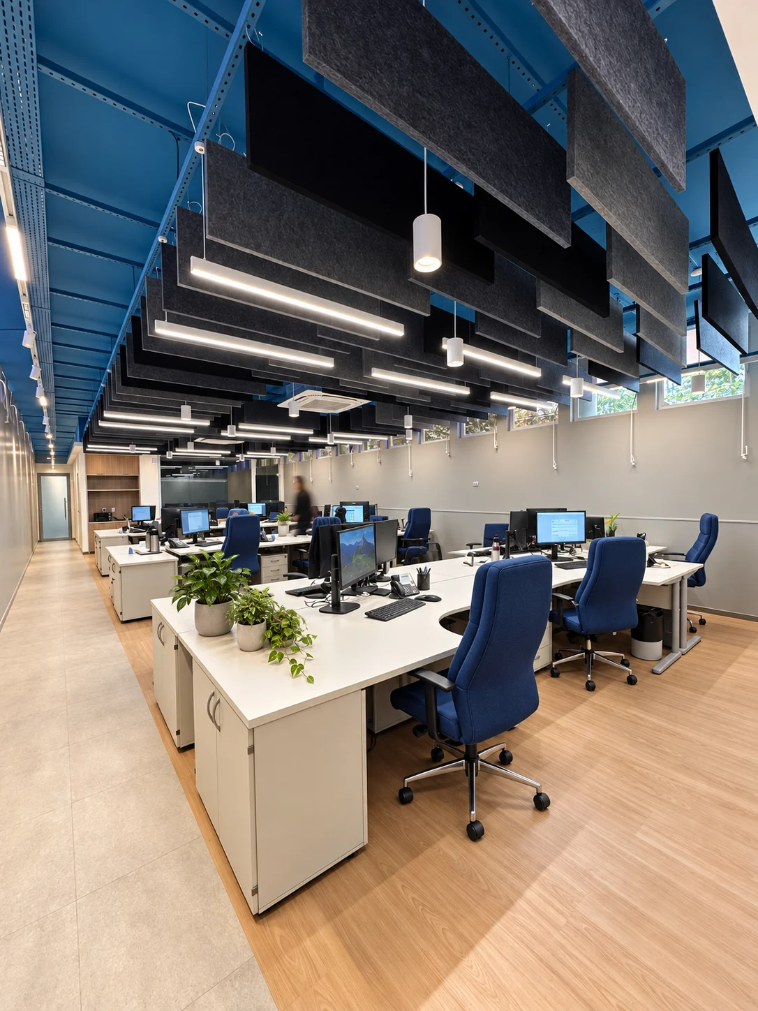 Open space da sede: bancadas contínuas com estações de trabalho, cadeiras azuis, forro em baffles acústicos escuros e estrutura metálica azul aparente