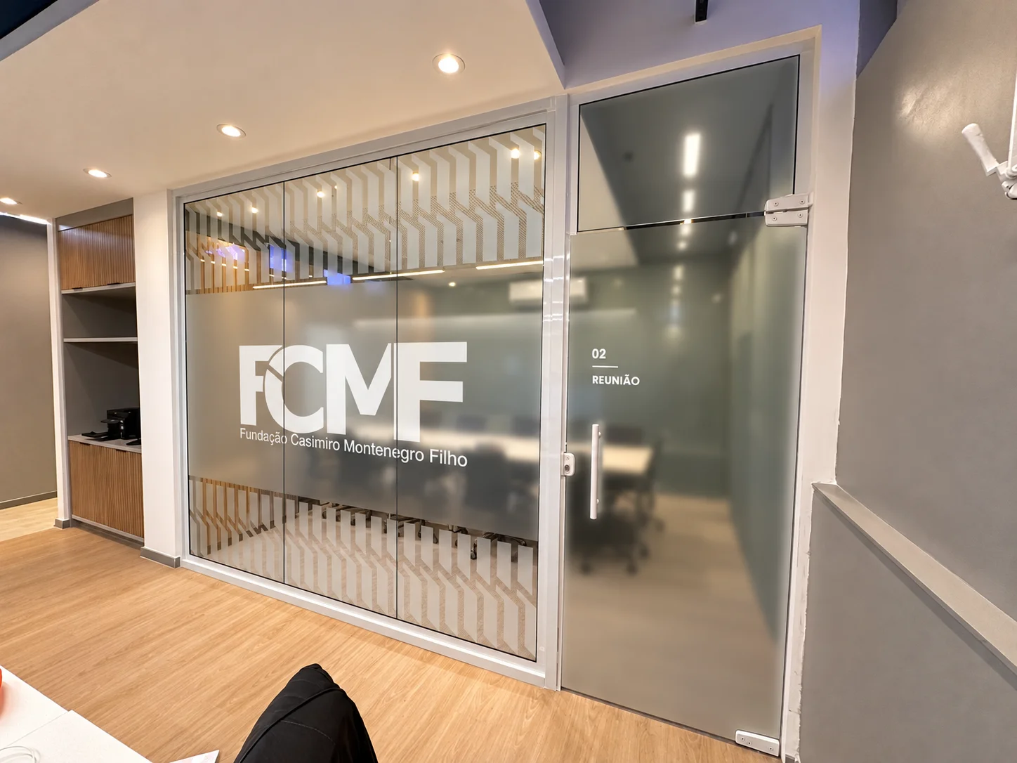 Divisória envidraçada da sala de reunião 02, com a marca da FCMF em película e o interior visível ao fundo