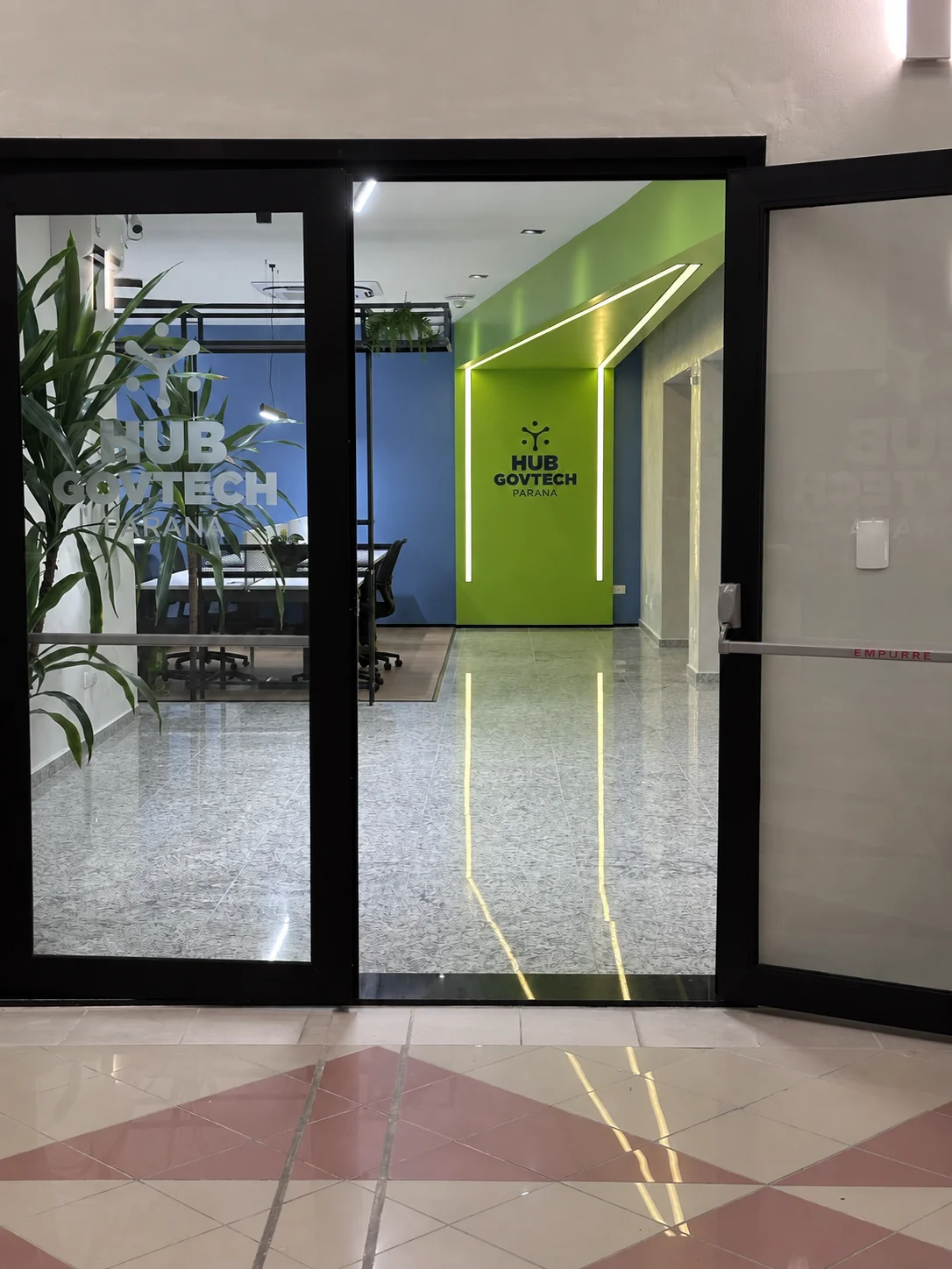 Acesso principal do hub: portas de vidro com a marca aplicada e a parede verde iluminada ao fundo