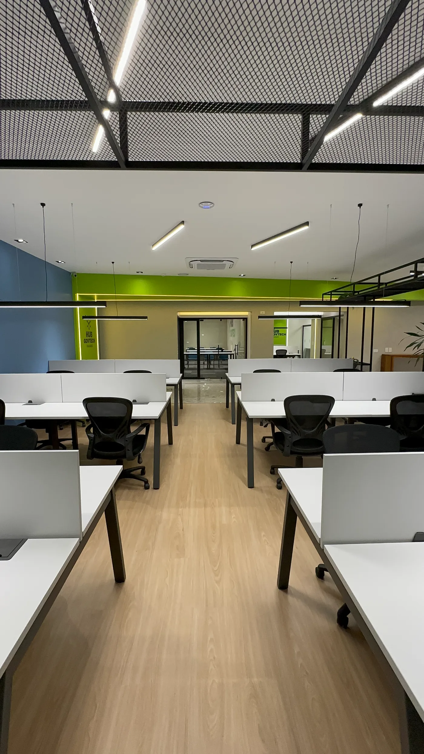 Eixo central do coworking, com as fileiras de estações voltadas para as salas envidraçadas e a marca aplicada na parede verde