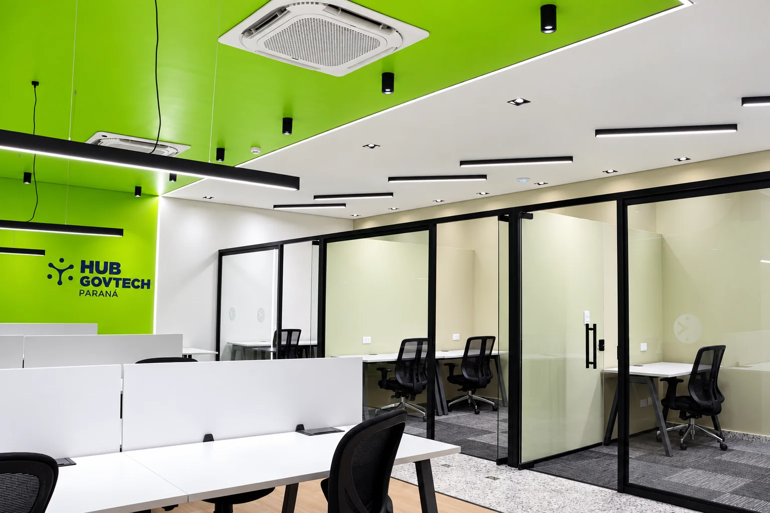 Estações de trabalho diante da parede verde com a marca do Hub GovTech Paraná e, ao lado, cabines individuais envidraçadas com caixilhos pretos