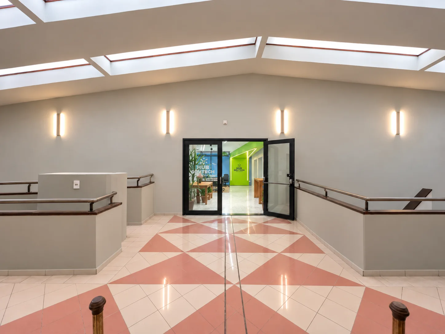 Hall principal requalificado: paredes em cinza, arandelas lineares, guarda-corpos com corrimão metálico e, ao fundo, a porta de vidro aberta para o Hub, com a marca na parede verde