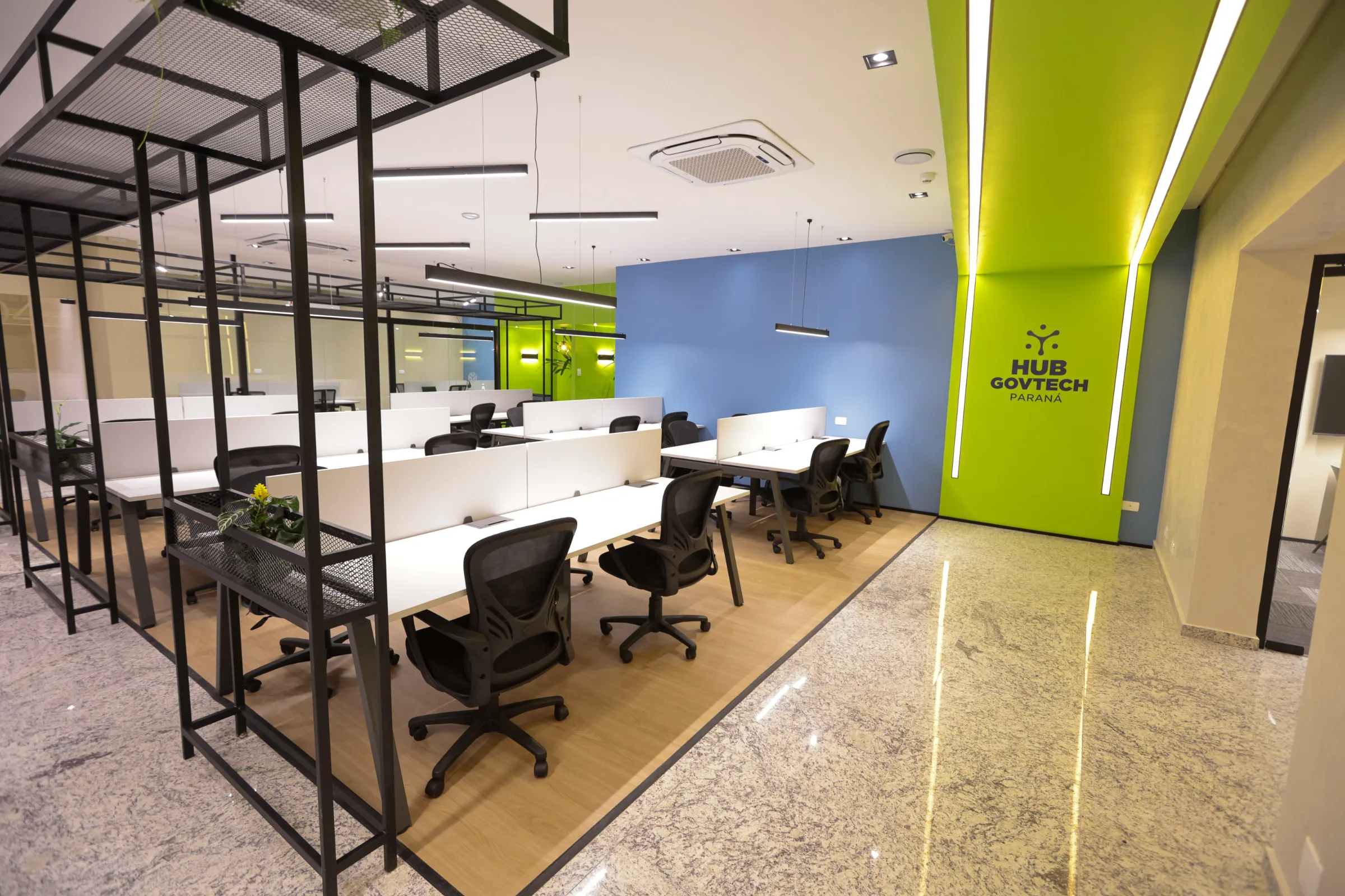 Hub GovTech Paraná: coworking com estações sobre piso de madeira, estruturas metálicas com vegetação e a marca do hub na parede verde