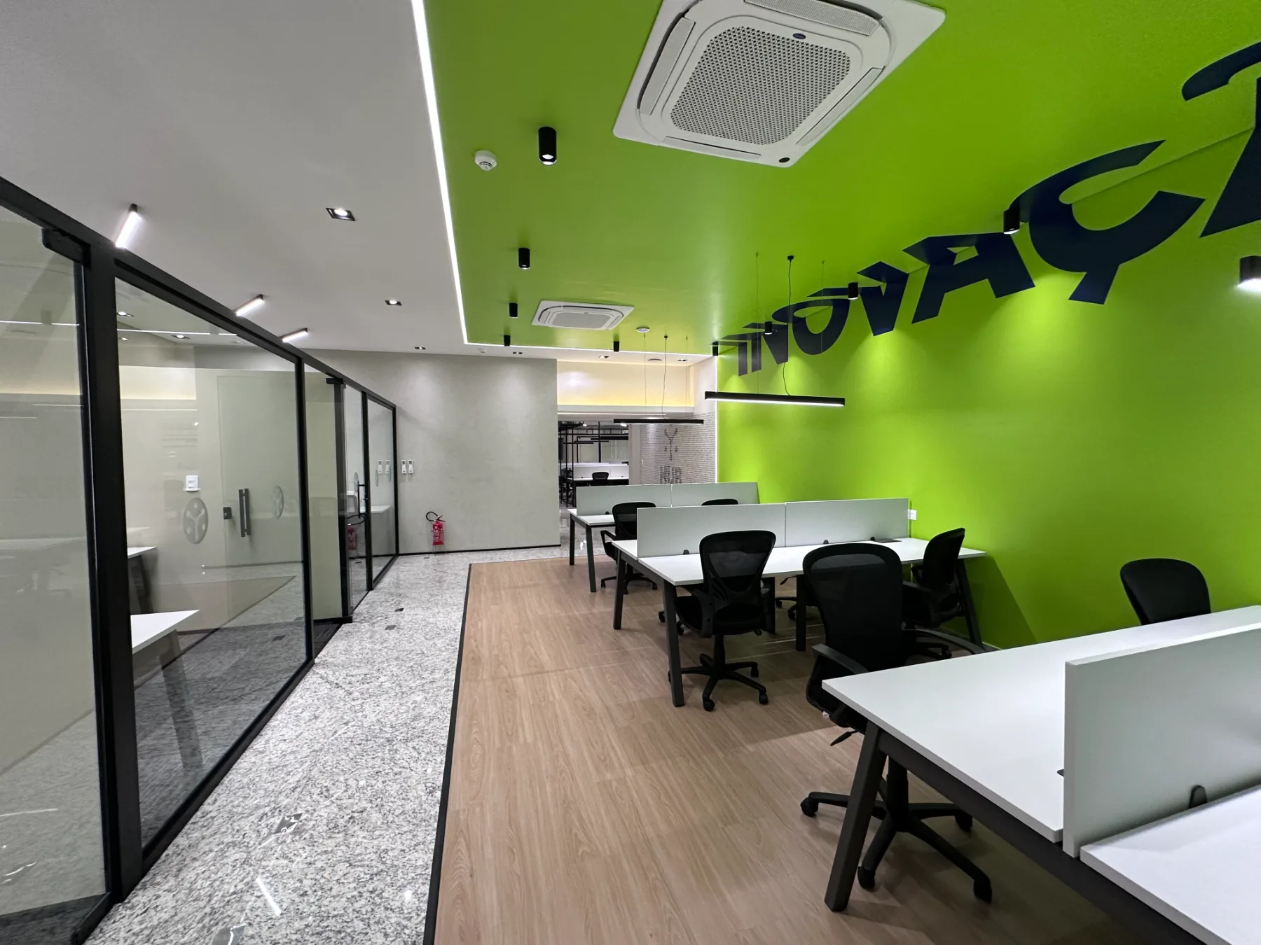 Estações de trabalho junto à parede verde com a palavra Inovação aplicada em tipografia recortada, e salas envidraçadas à esquerda