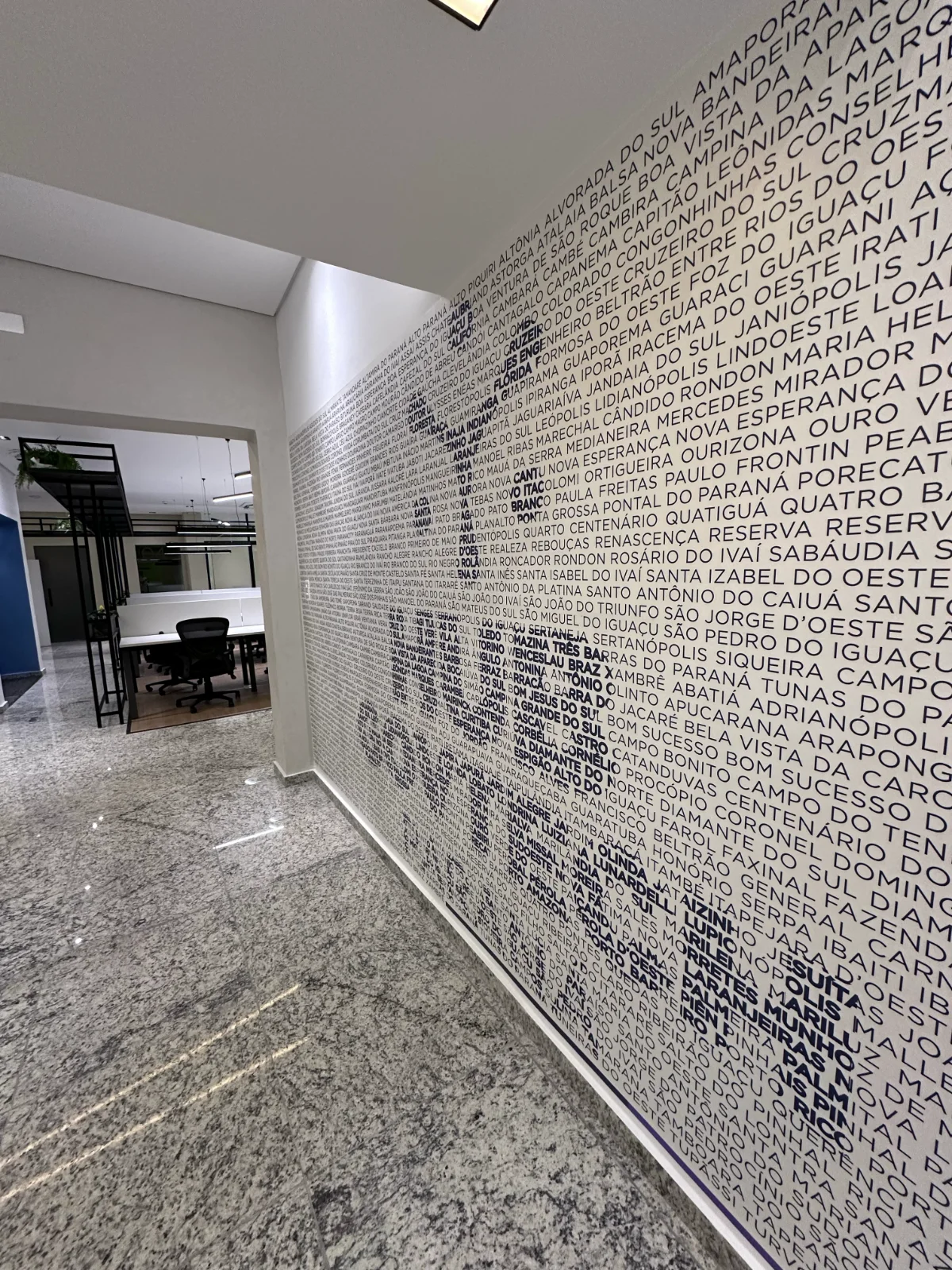 Parede da circulação revestida com os nomes dos municípios do Paraná em tipografia azul, formando o símbolo da marca por contraste de peso
