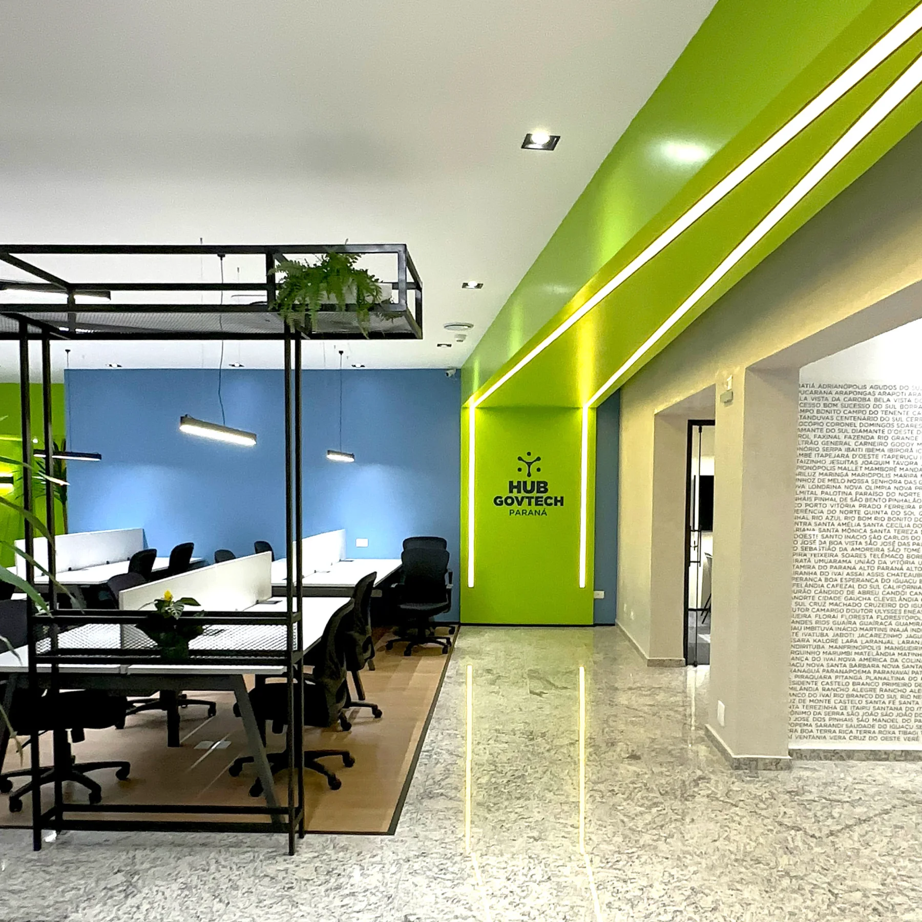 Vista geral do Hub GovTech: estações de trabalho sob estrutura metálica com vegetação, parede azul ao fundo, portal verde iluminado com a marca e a parede dos municípios do Paraná à direita