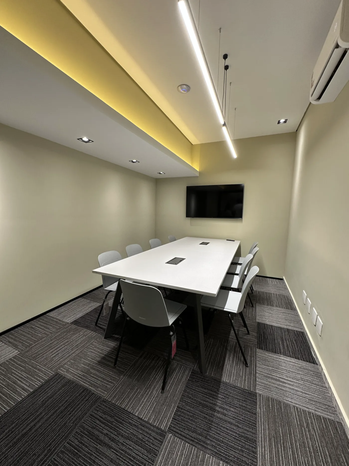 Sala de reunião para dez lugares, com sanca iluminada em amarelo, pendente linear e televisor na parede