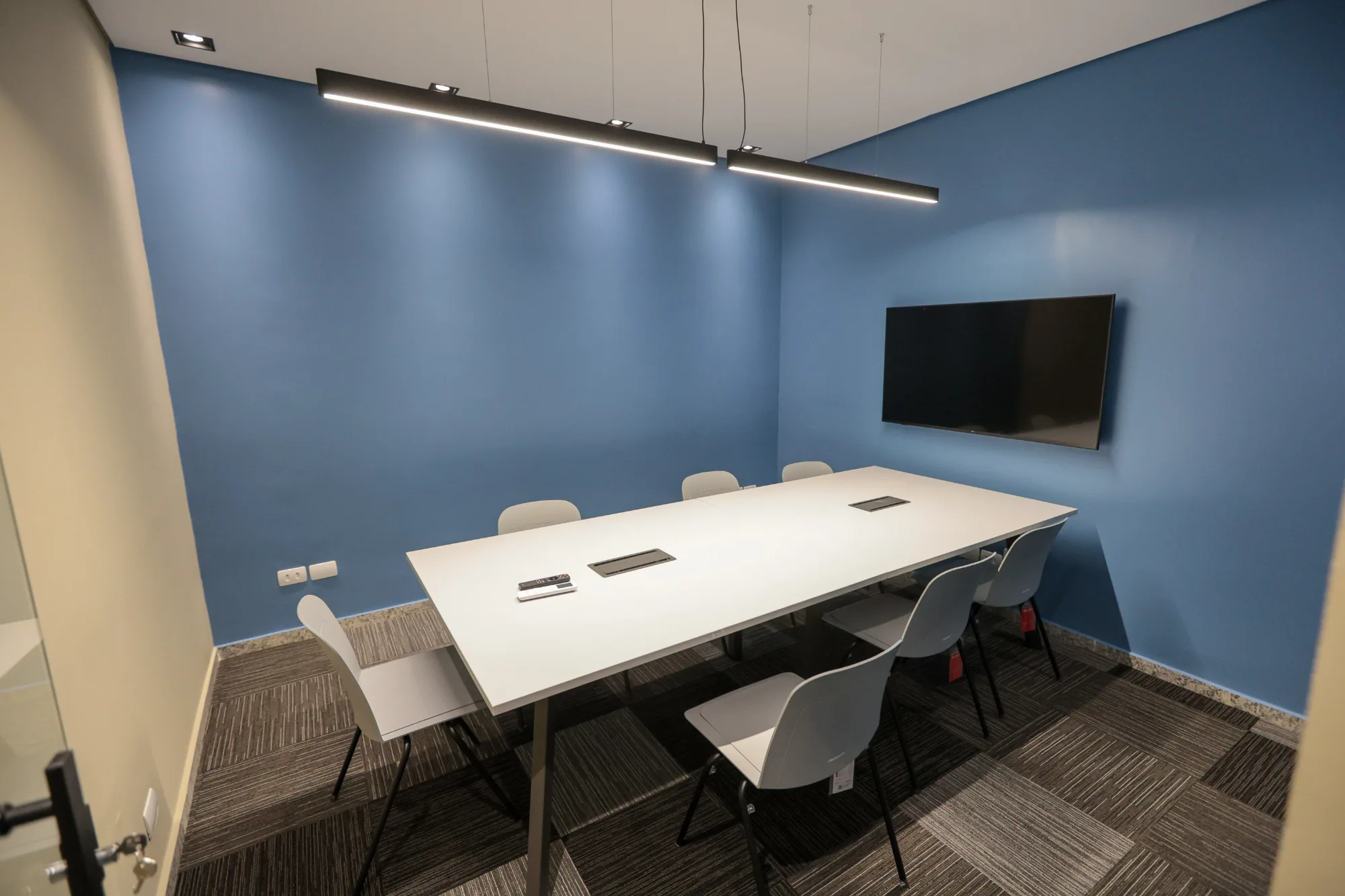 Sala de reunião com parede azul, mesa branca para oito lugares e pendente linear sobre a mesa