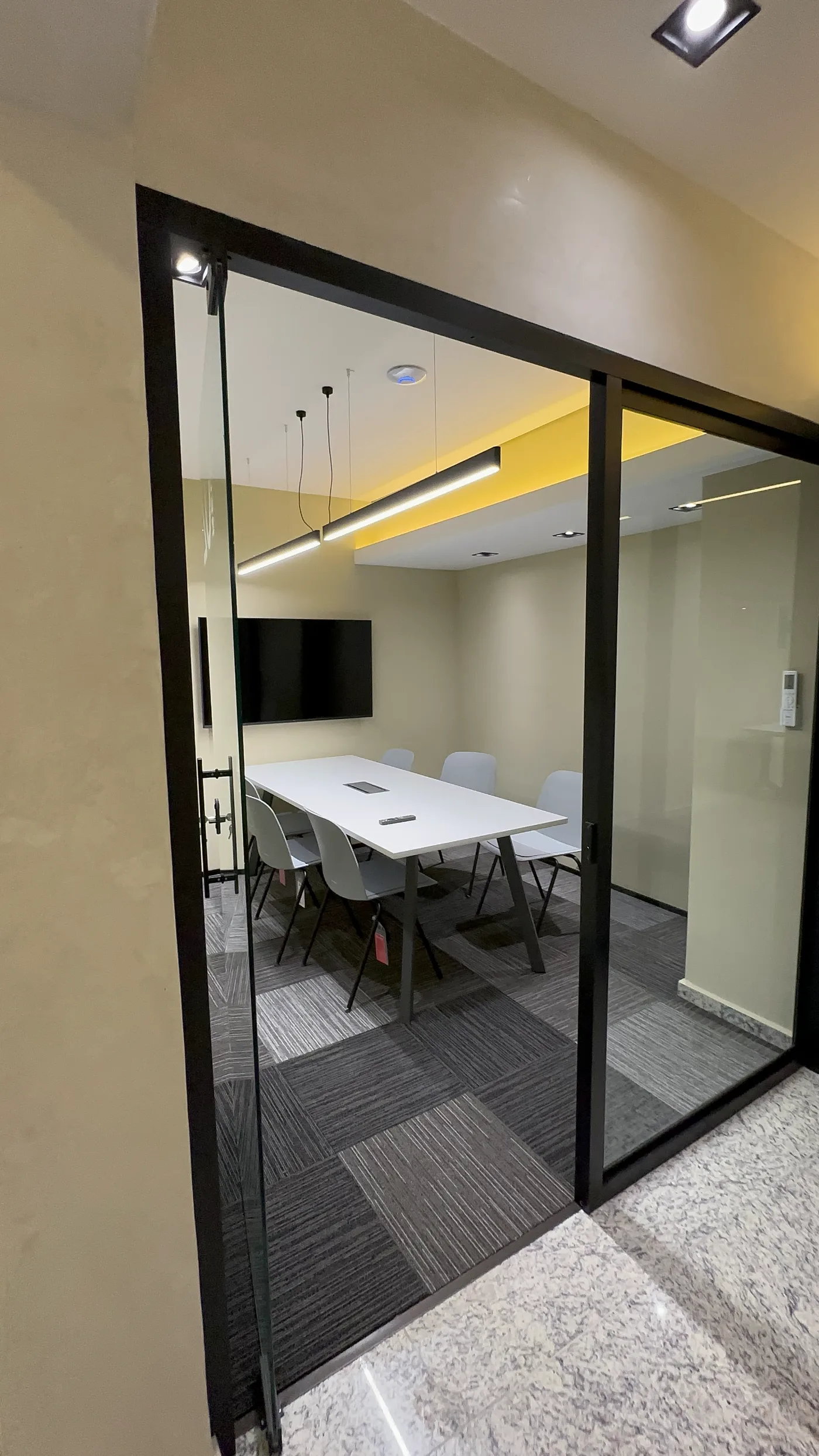 Sala de reunião envidraçada vista da circulação, com mesa branca e parede amarela sob o forro rebaixado