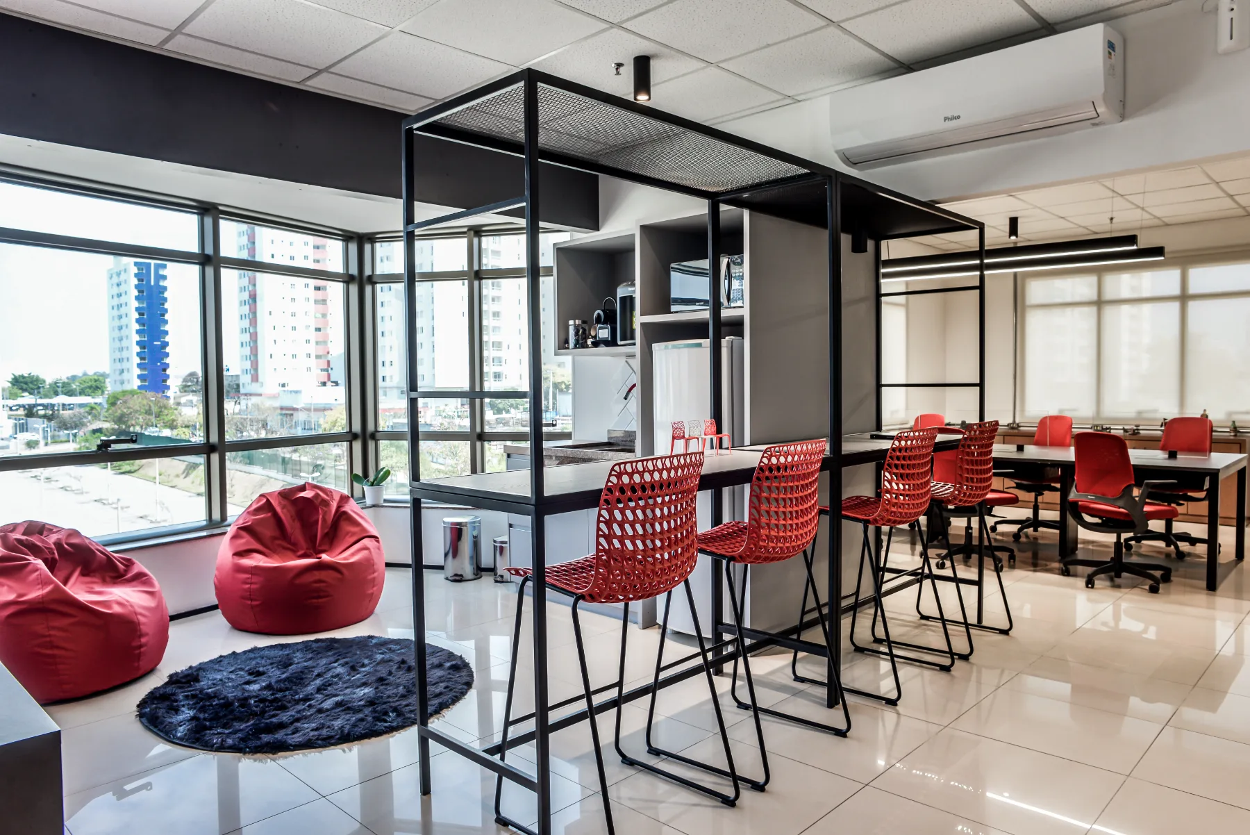 Área de convivência do Grupo Txai: pórtico metálico com bancada alta e banquetas vermelhas, pufes junto à janela e estações de trabalho ao fundo