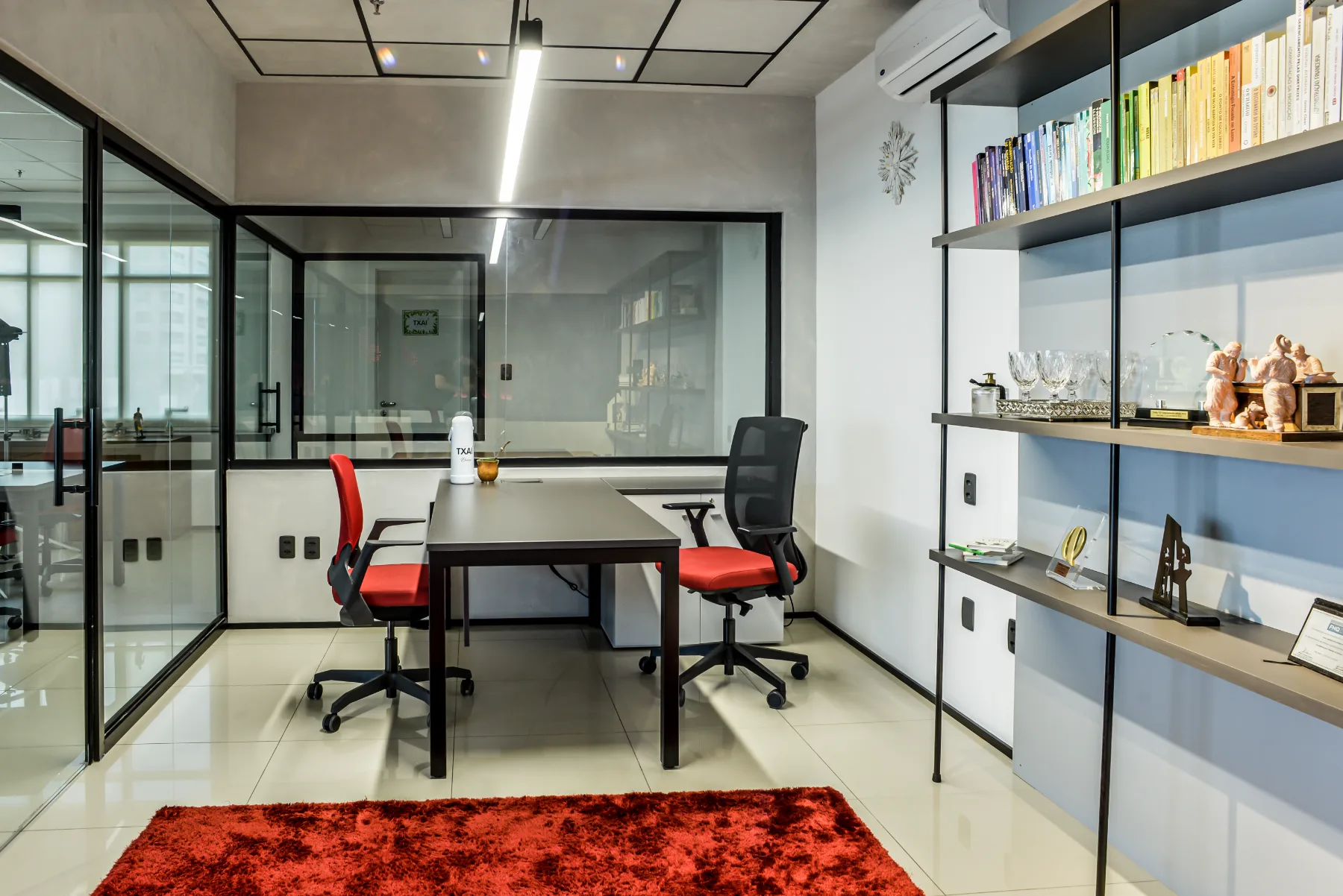 Sala de gestão com mesa em L, estante metálica de livros e reconhecimentos, e divisórias de vidro para a operação