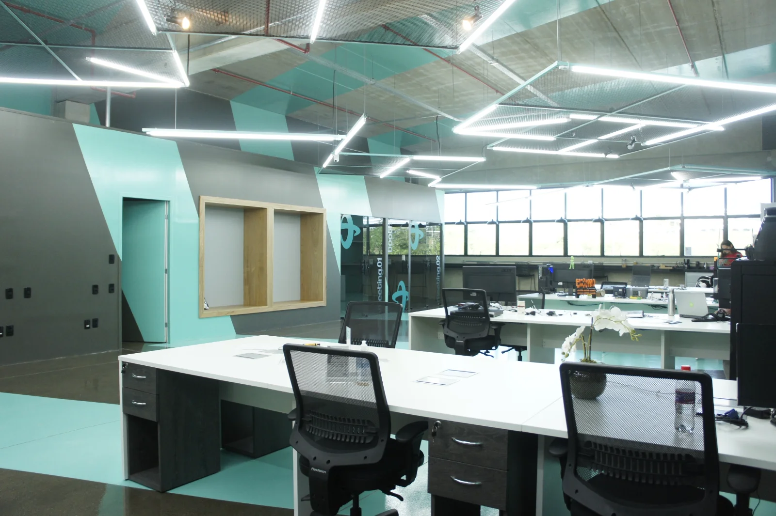 Estações de trabalho compartilhadas em open space, com armários baixos e phone booths envidraçados ao fundo