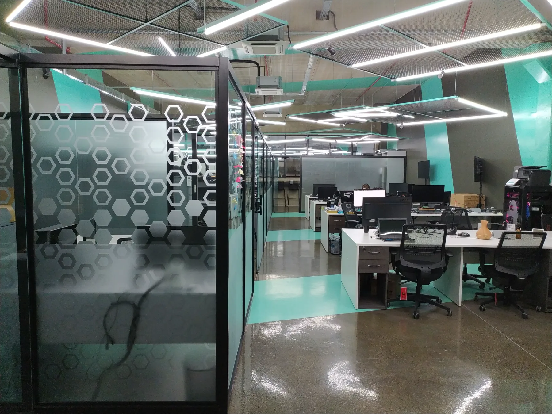 Divisórias de vidro com grafismo hexagonal separando salas e a faixa contínua de estações de trabalho