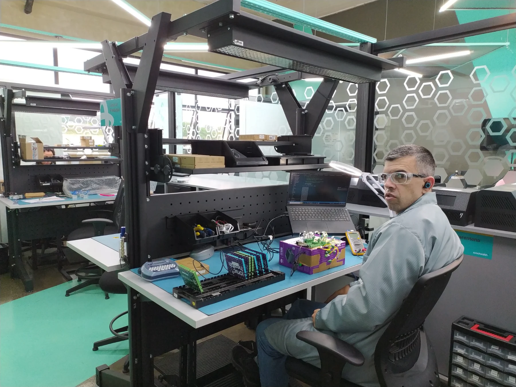 Técnico em bancada antiestática da área de produção, com iluminação dedicada e divisória de vidro com grafismo hexagonal ao fundo