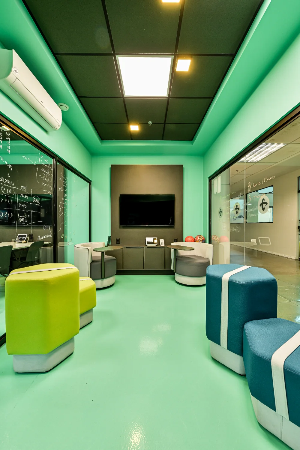 Sala de descompressão com pufes coloridos, sofá e televisor, integralmente revestida em verde-água