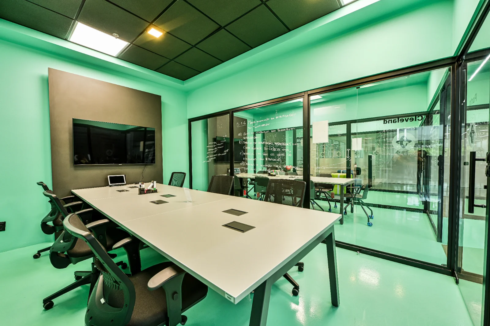 Sala de reunião com mesa longa, televisor e divisórias de vidro voltadas para a operação