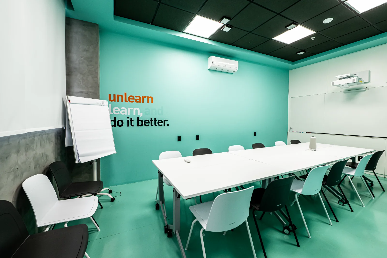 Sala de treinamento com mesas em U, flip chart e a frase unlearn, learn, and do it better na parede
