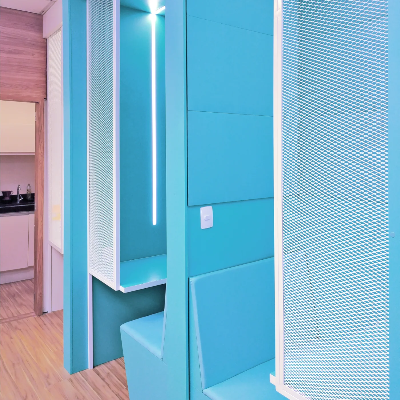 Phone booth construído em turquesa com bancada, assento estofado, iluminação linear e fechamento em tela metálica branca
