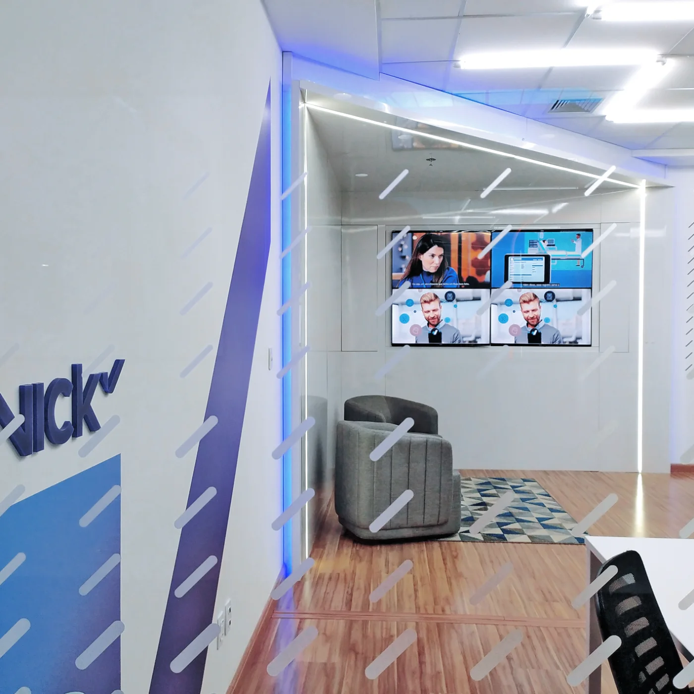 Sala de apresentação com videowall de quatro telas, poltronas cinza e marca em relevo na parede com grafismo aplicado