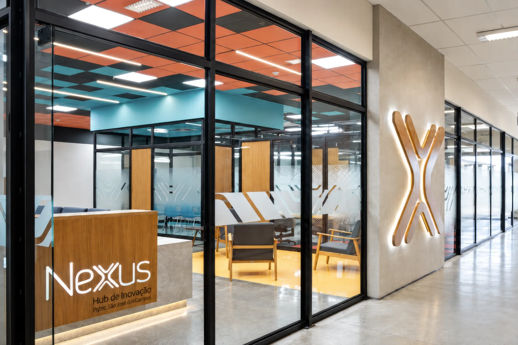 Entrada do Coworking Júpiter: recepção em madeira atrás do vidro, forro em xadrez colorido e marca retroiluminada na parede da circulação