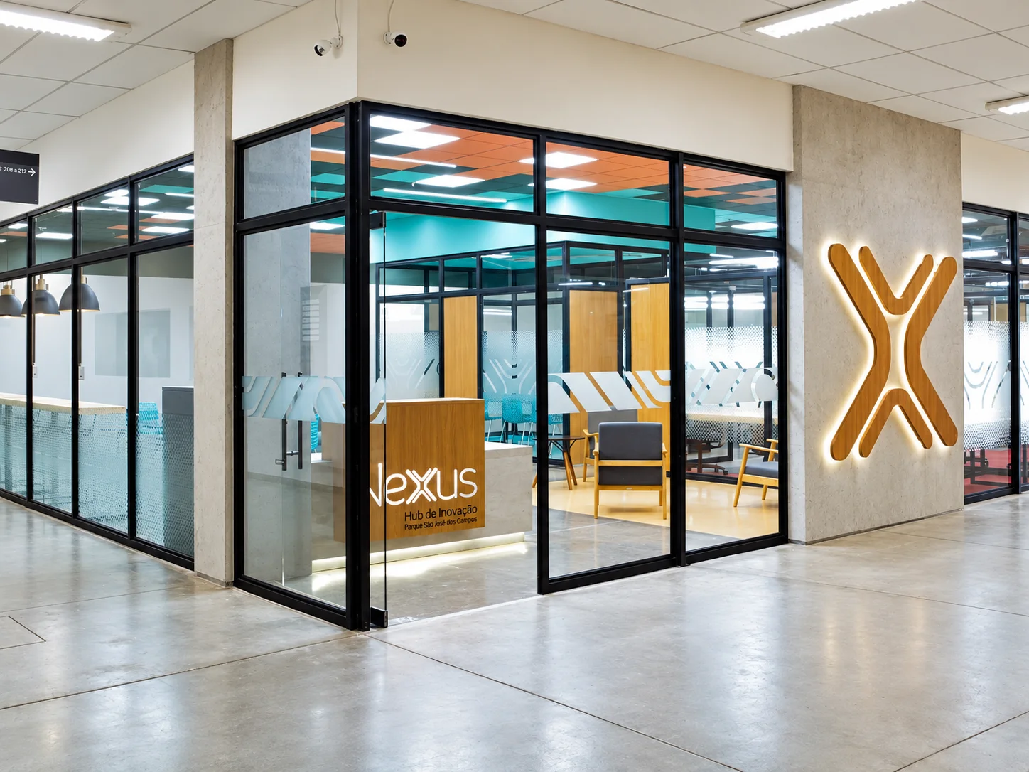 Entrada do coworking na esquina do corredor, com fachada envidraçada em caixilhos pretos, balcão de recepção em madeira com a marca Nexus e o símbolo X iluminado na parede de concreto