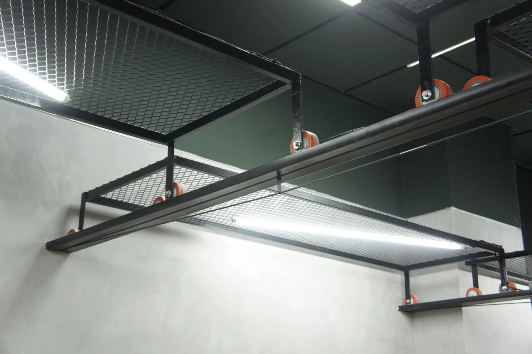 Detalhe das bandejas metálicas suspensas em tela expandida, com rodízios aparentes