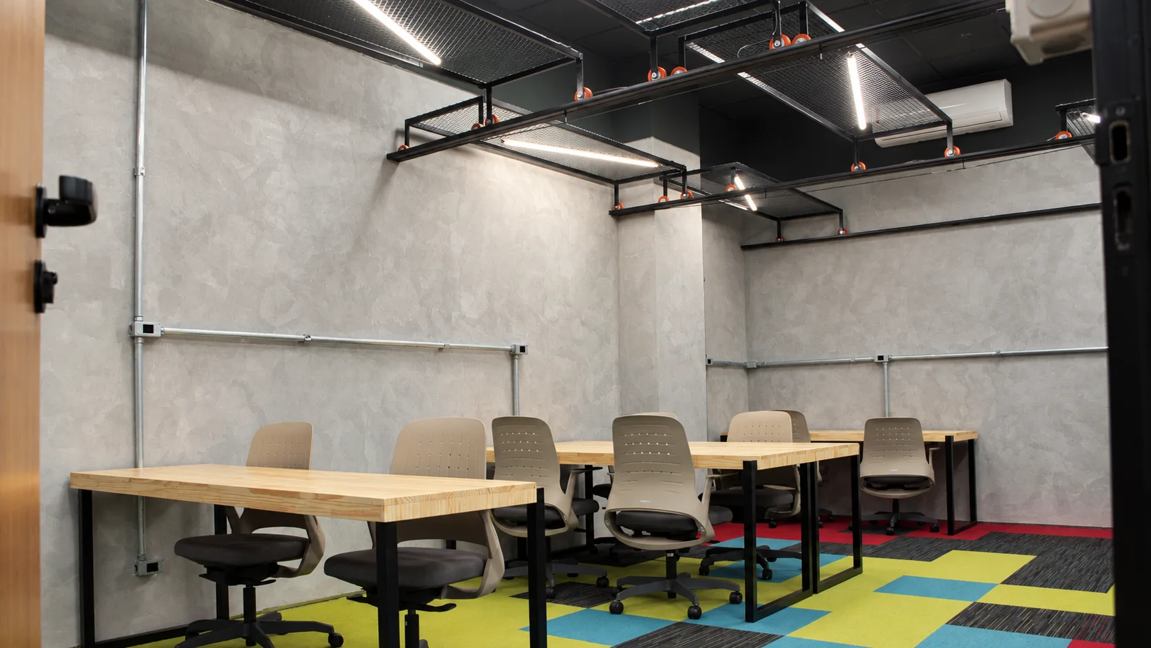 Sala privativa com paredes em cimento queimado, bandejas metálicas suspensas, bancadas de madeira e carpete modular colorido em verde, azul, vermelho e cinza