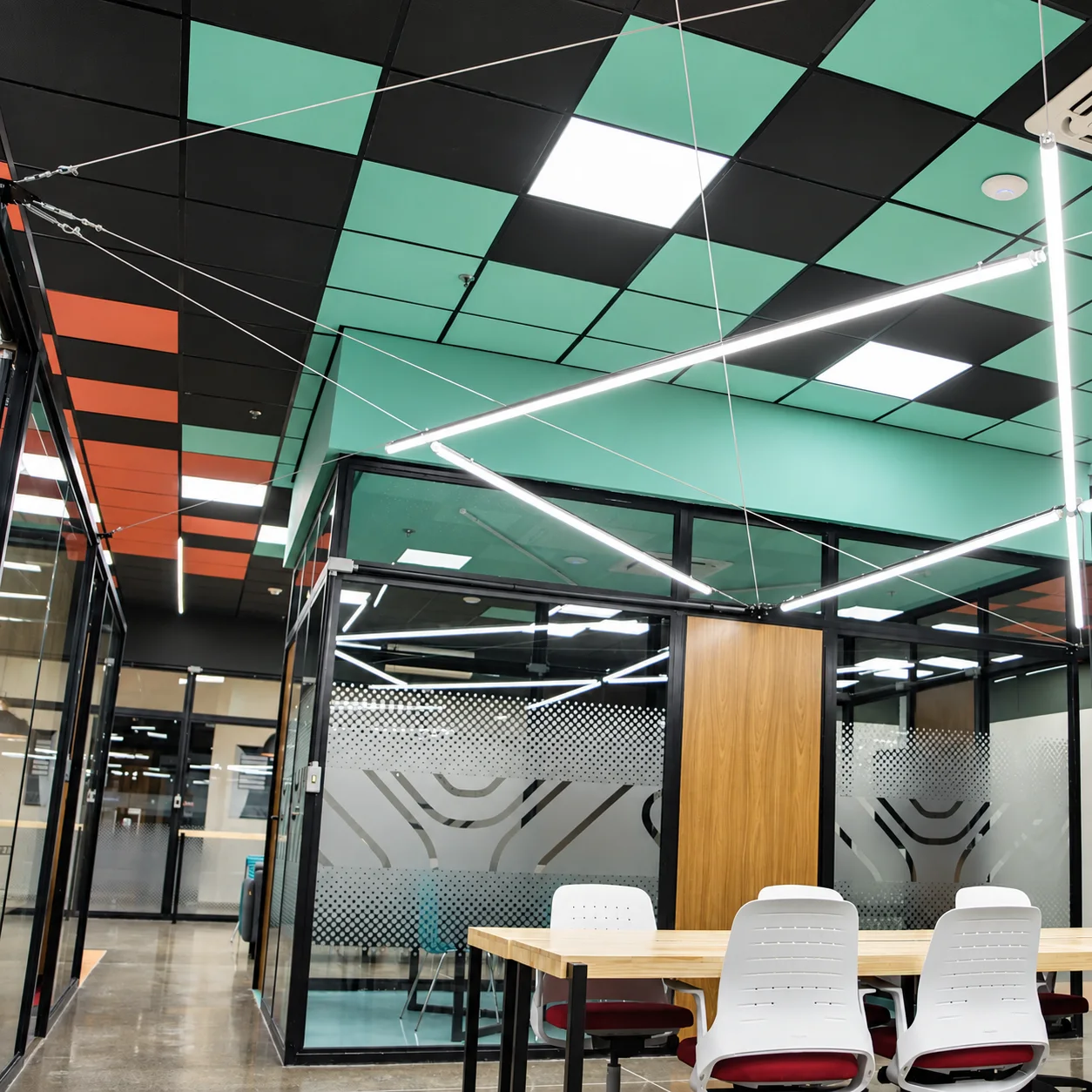 Sala de reunião envidraçada com película de padrão pontilhado e grafismo do símbolo, sob o forro em xadrez verde-água e preto, com luminárias lineares suspensas por cabos e mesa de madeira em primeiro plano