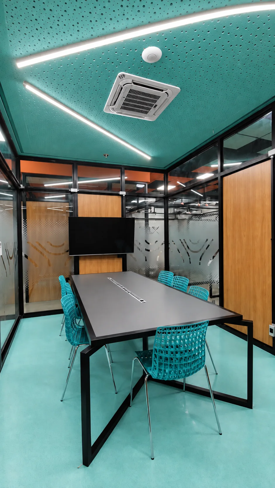 Sala de reunião em turquesa, com forro acústico perfurado, piso na mesma cor, divisórias em vidro e madeira, mesa preta e cadeiras vazadas turquesa