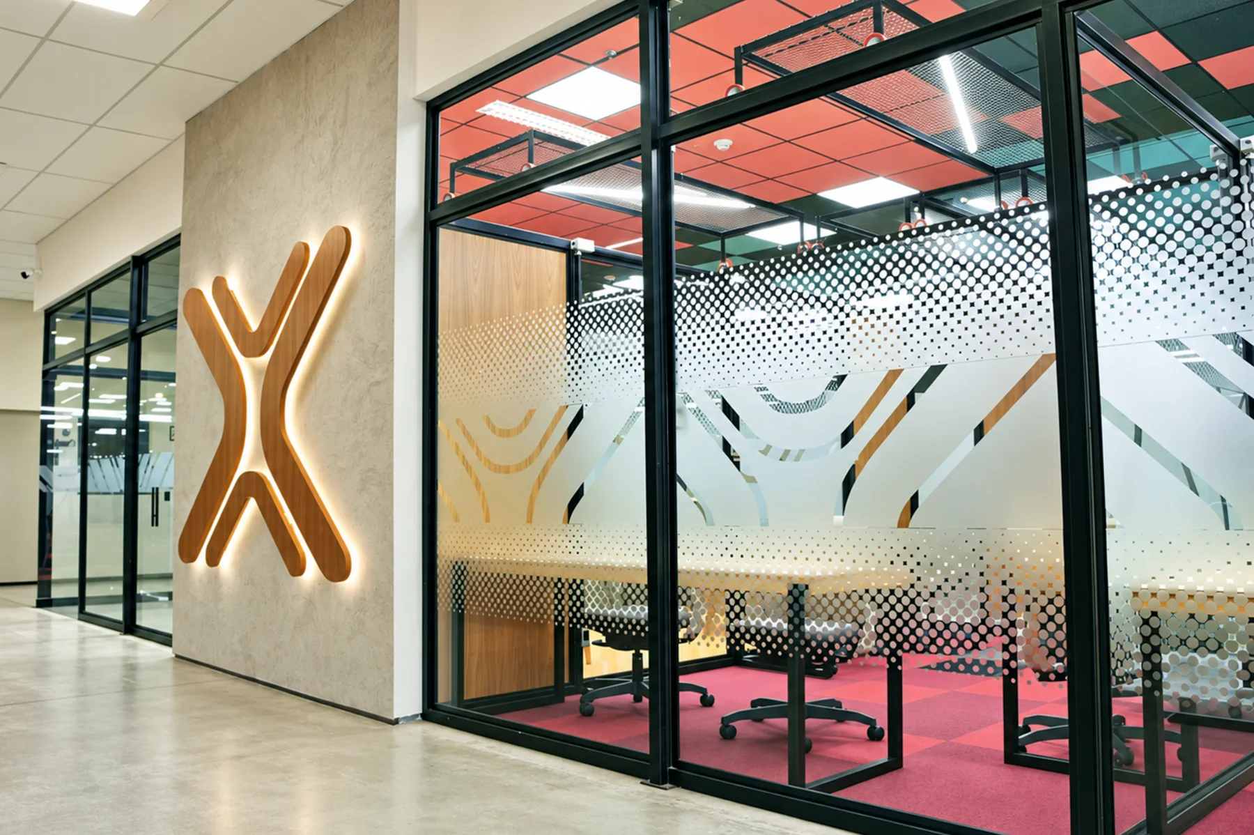 Marca X em madeira retroiluminada na parede de chegada e, ao lado, sala envidraçada com película de padrão pontilhado e grafismo derivado do símbolo