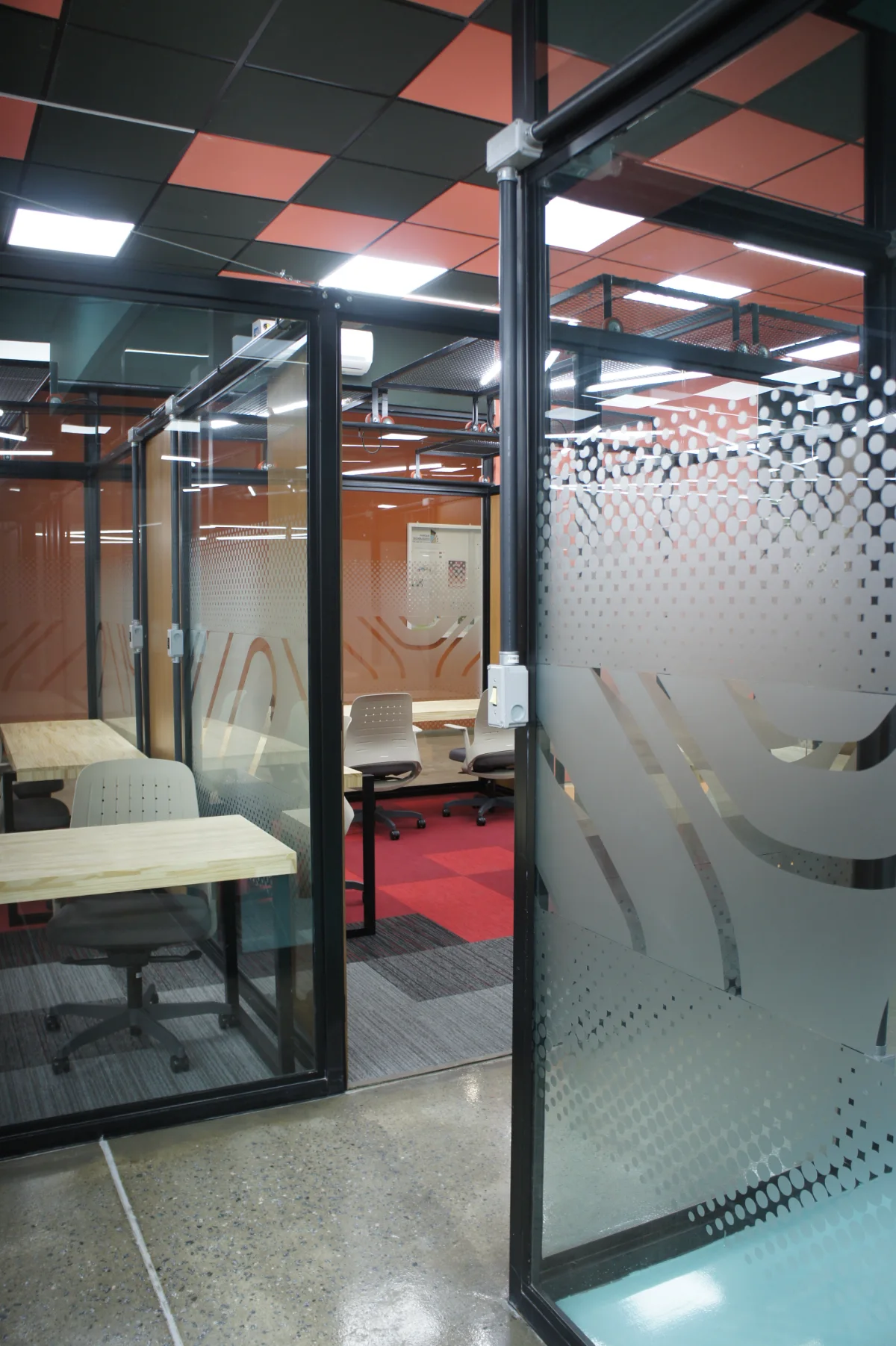 Porta de vidro com película de padrão pontilhado e grafismo derivado do símbolo, abrindo para salas privativas com carpete modular vermelho e forro em xadrez coral e preto
