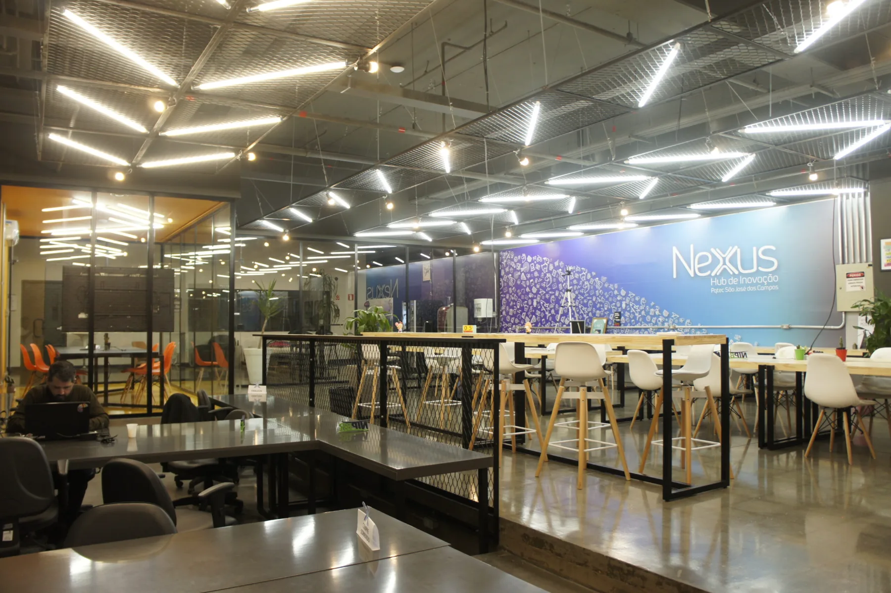 Salão principal do Coworking Netuno, com bancadas compartilhadas, forro em tela expandida iluminada e a marca aplicada na parede ao fundo