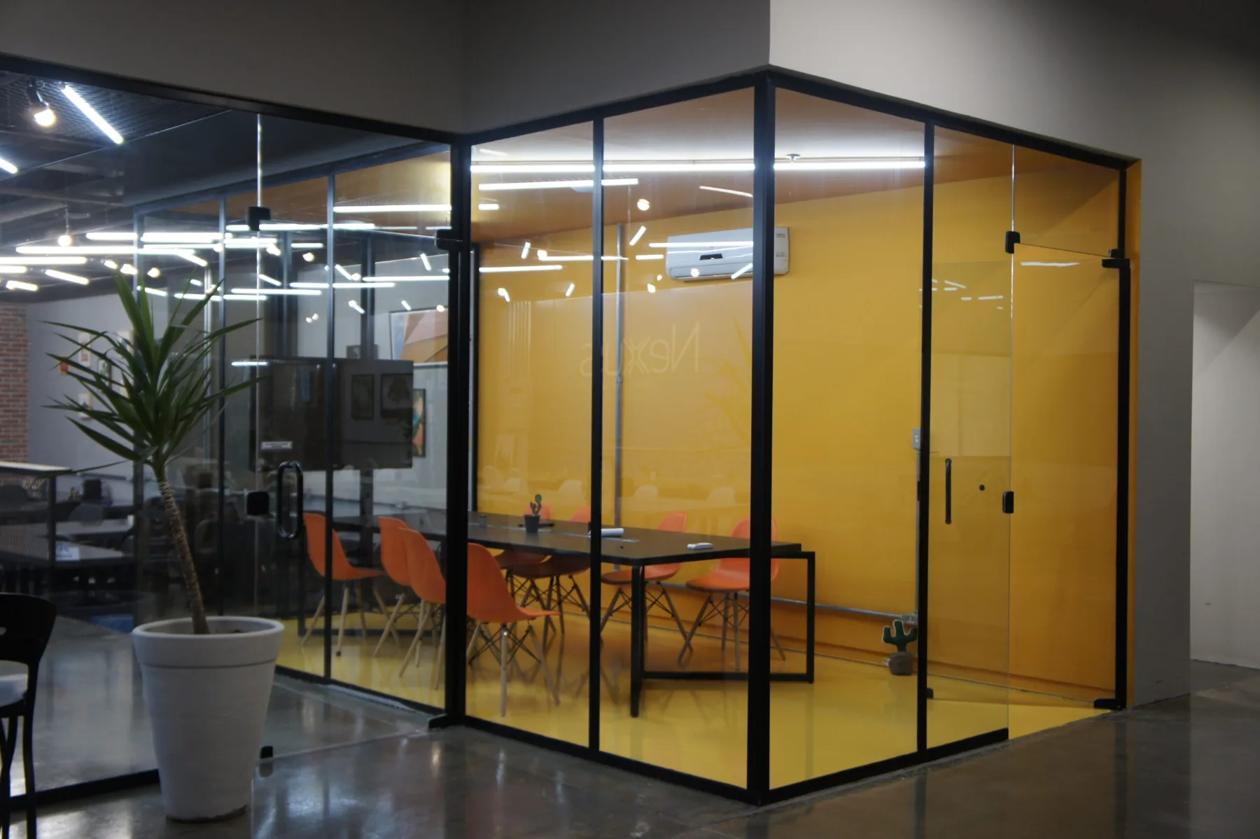 Sala de reunião envidraçada com piso e parede amarelos, mesa longa e cadeiras laranja