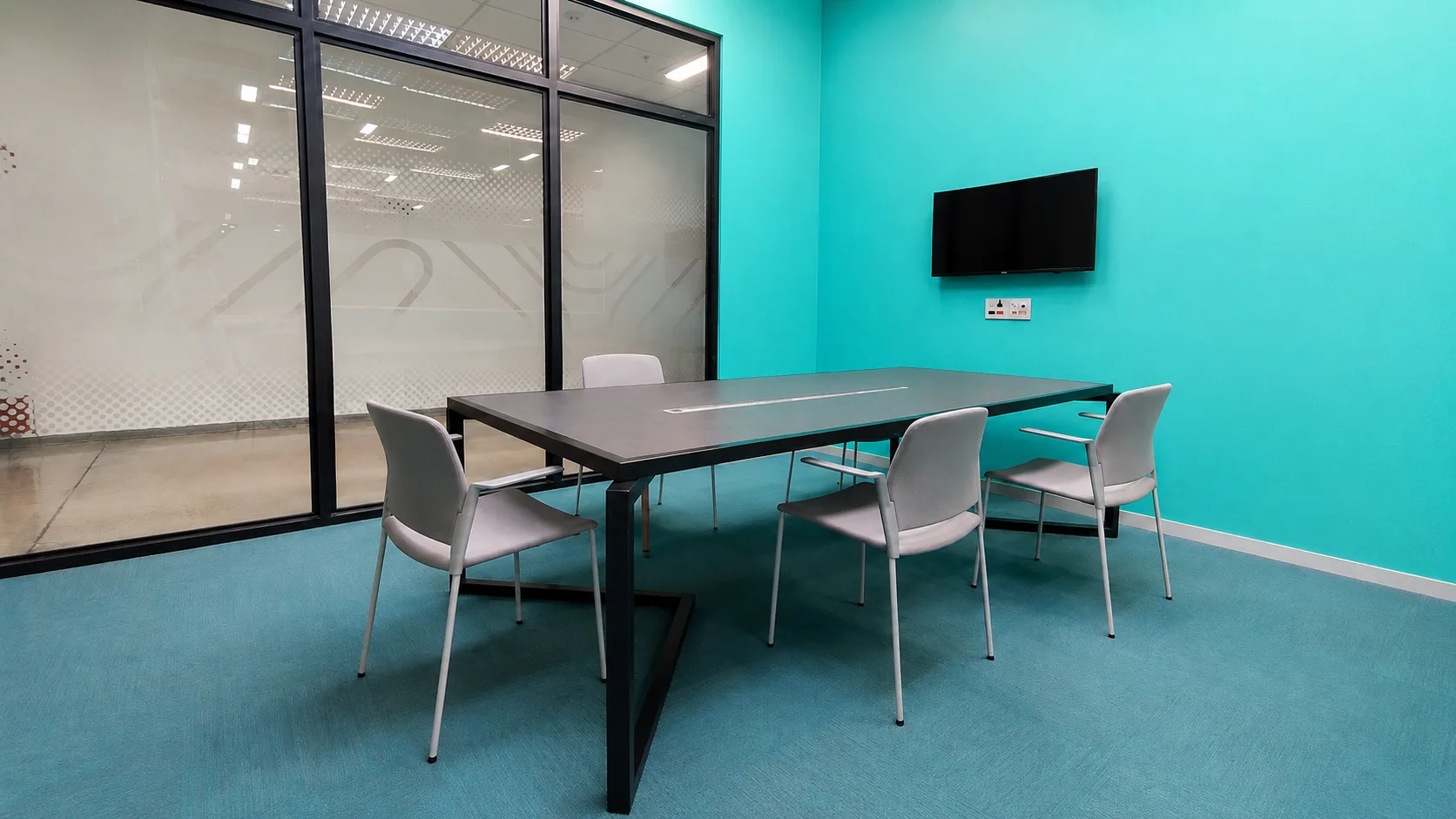 Sala de reunião com parede turquesa, carpete azul, mesa escura e monitor de parede