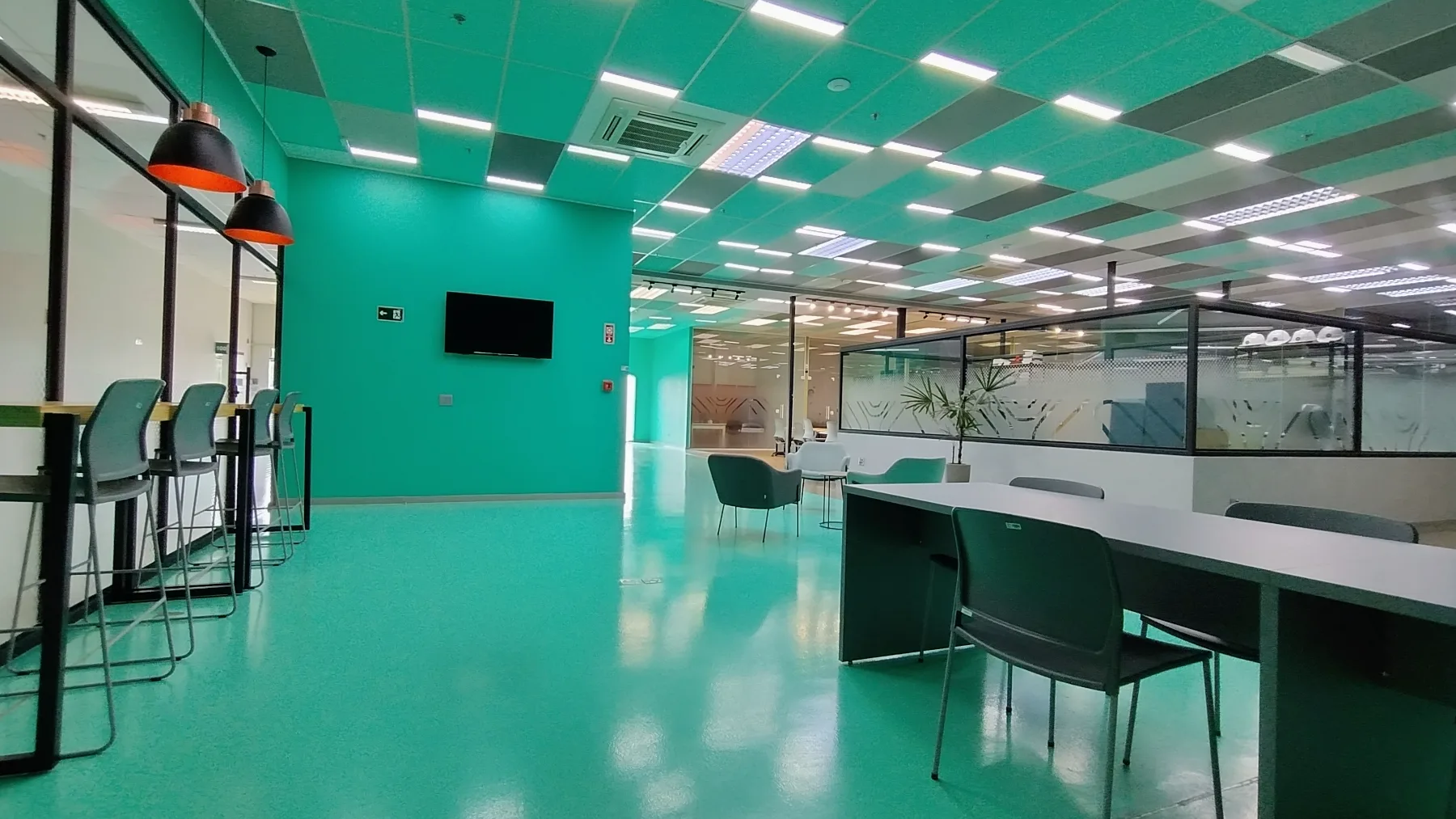 Área ampla do Coworking Urano, com piso em resina turquesa, bancadas de apoio e salas envidraçadas ao fundo