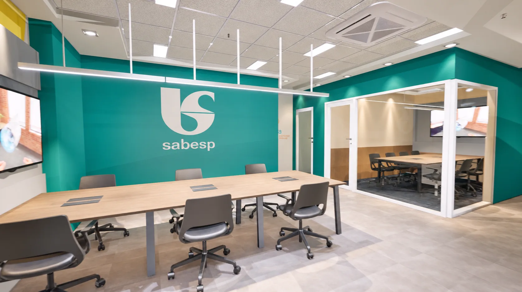 Área de trabalho compartilhada diante da parede turquesa com a marca SABESP, e sala de reunião envidraçada ao lado