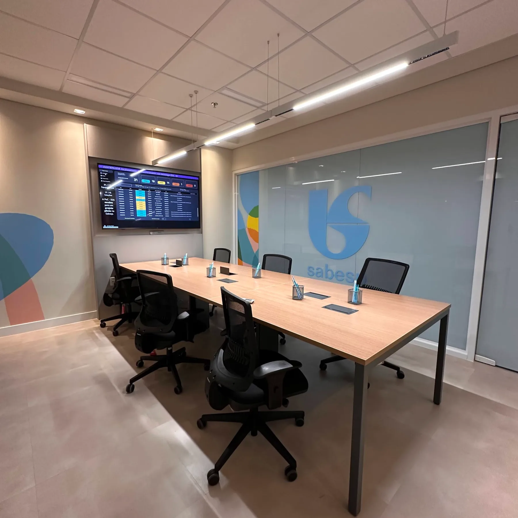 Sala de reunião principal, com mesa longa, tela de monitoramento e marca aplicada no vidro jateado