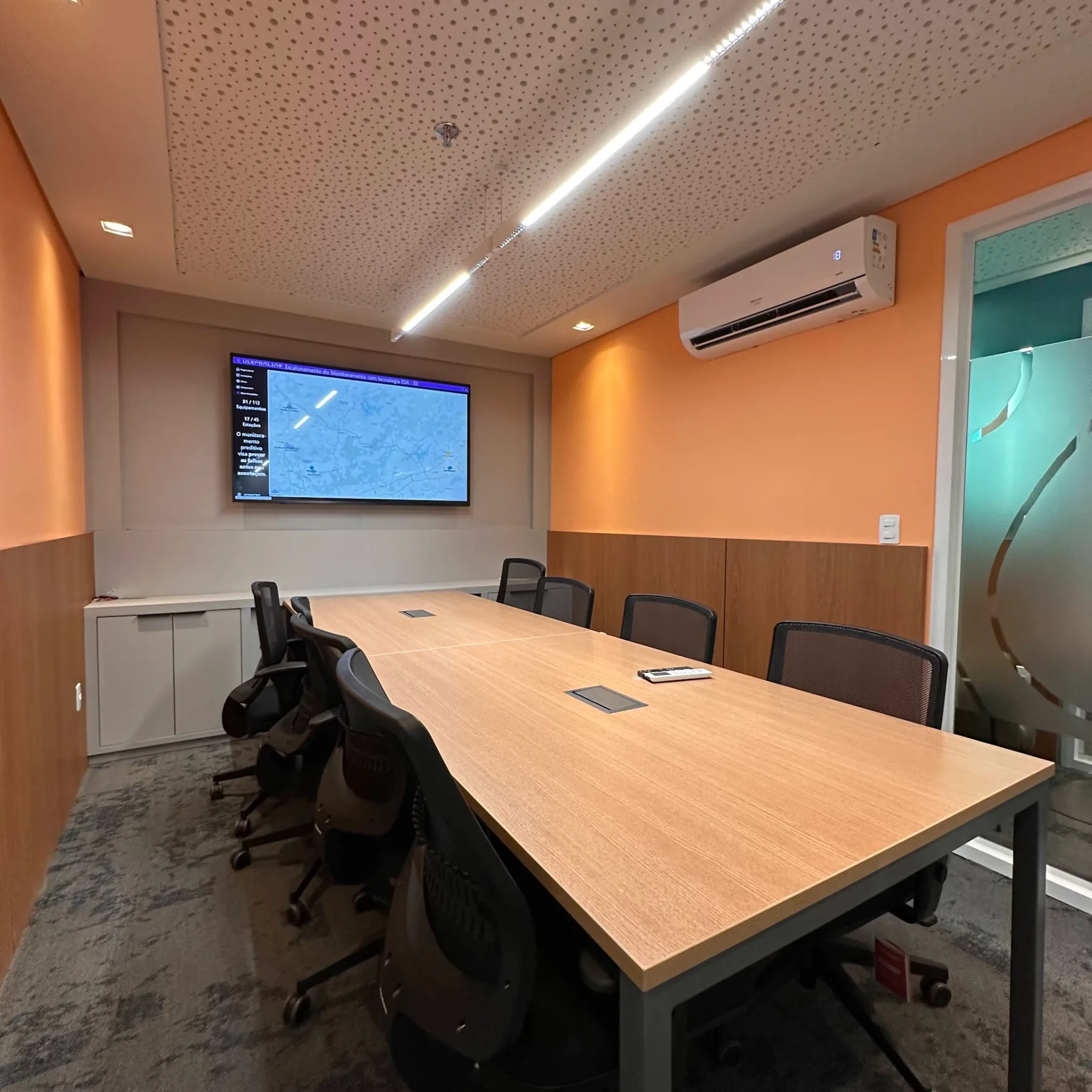 Sala de reunião com parede em tom laranja, marcenaria em madeira e tela de monitoramento
