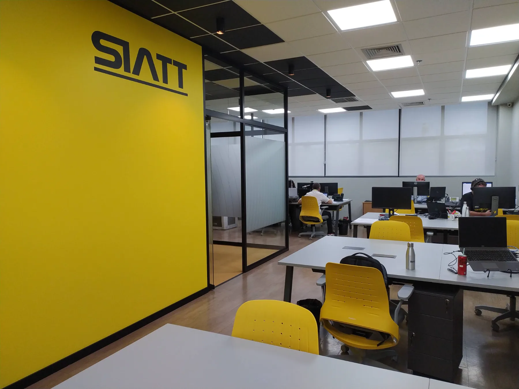 Parede amarela com a marca aplicada junto à sala envidraçada, com as estações de trabalho da operação à direita